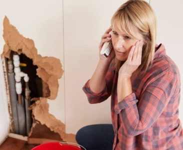 woman with blonde hair on the phone kneeling on wood floor, red bucket, burst pipe in white wall